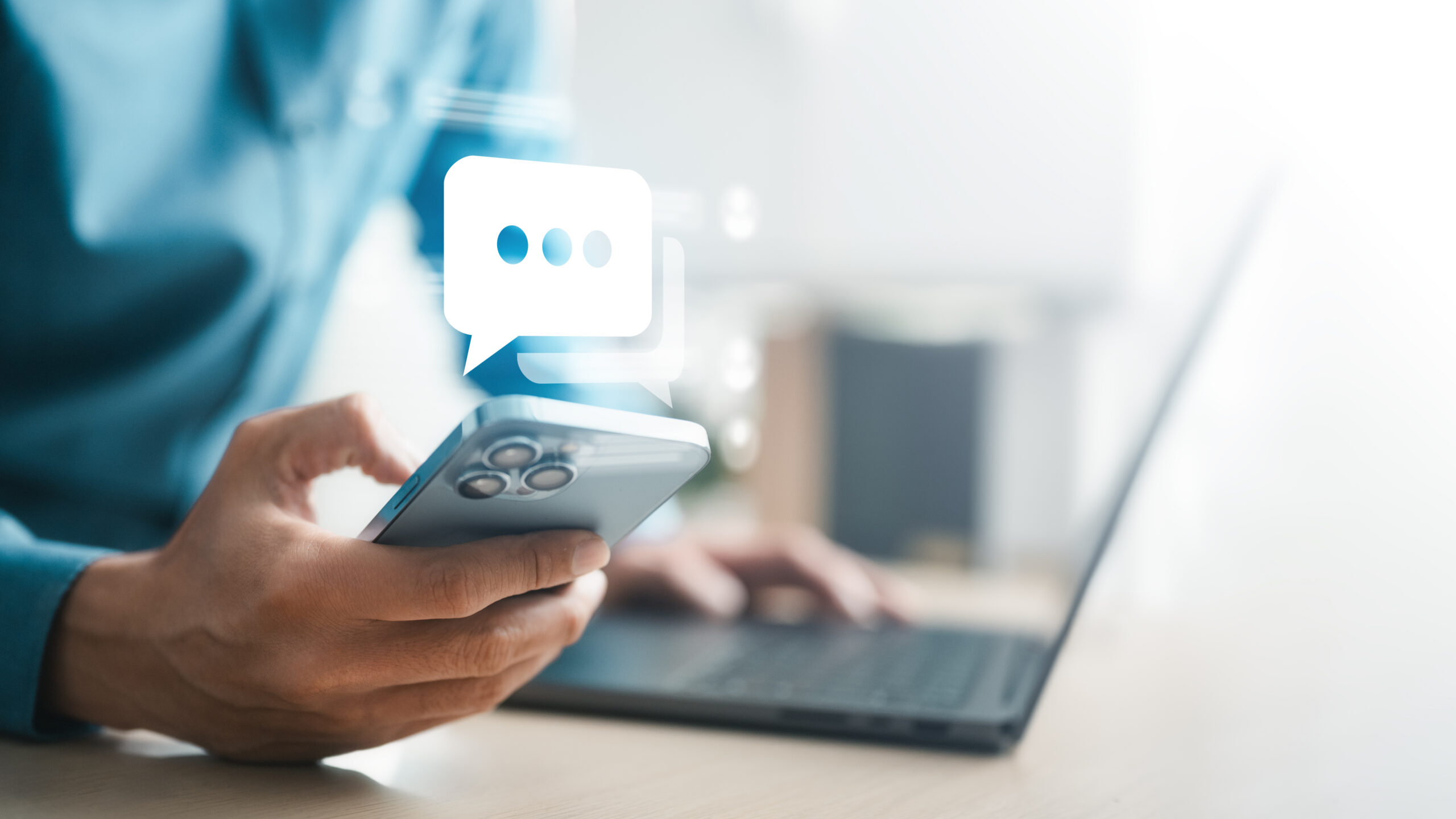 Person using smartphone beside laptop with chat icon overlay, representing online messaging, mobile communication, customer support, social media interaction, app notification, and digital connection.